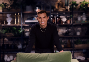 Matt Fraser, a man with short brown hair, wearing a black shirt and a gold chain necklace, stands behind a green couch in a room with shelves and plants in the background.